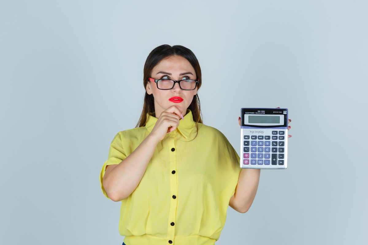 Mulher branca de óculos, com cara de dúvida, segurando uma calculadora para calcular a nota do enem.