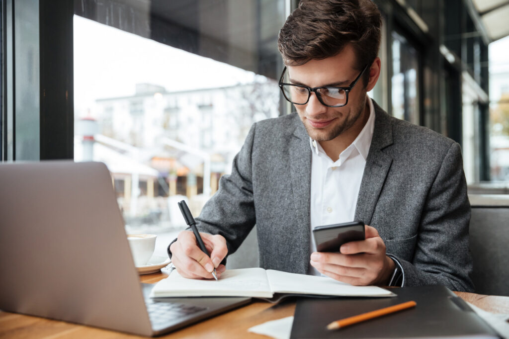 Profissional de administração sorridente de óculos, sentado à mesa no café com o laptop enquanto usa o smartphone e escreve algo.