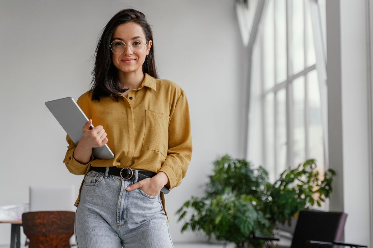 Mulher branca de cabelo preto e liso, em pé, com a mão direita no bolso da calça jeans. Ela segura uma pasta com a outra mão, com currículo profissional