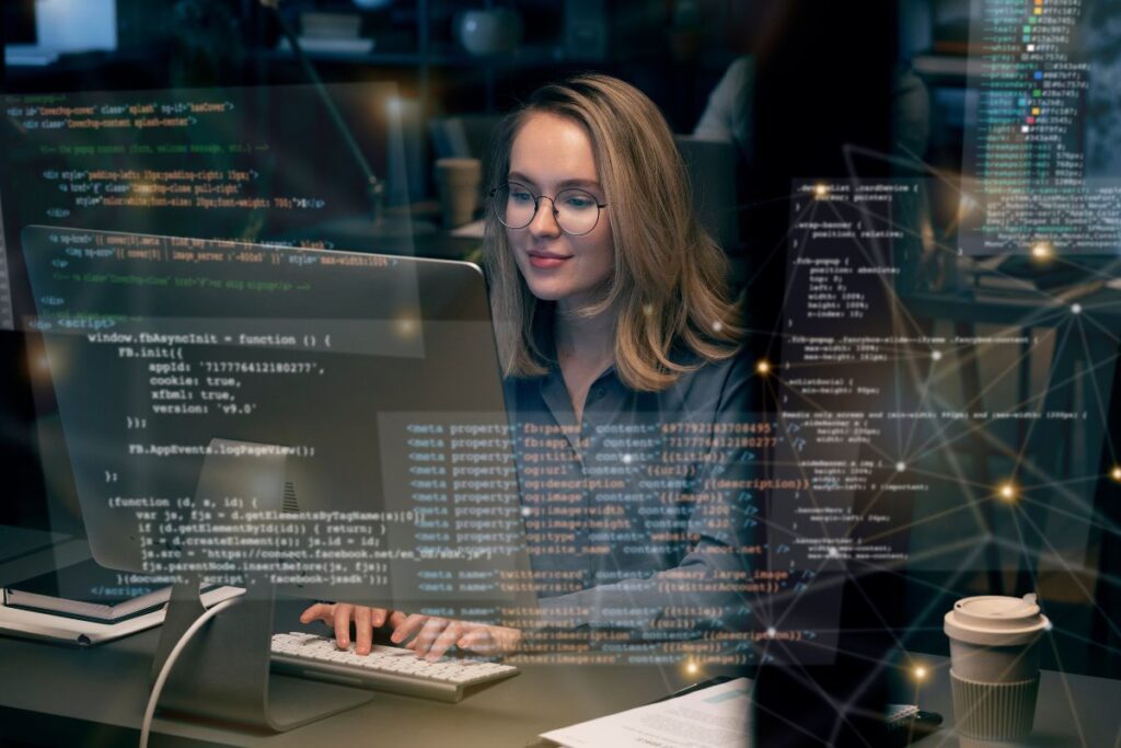 Uma das mulheres na tecnologia, branca, loira, de óculos, sentada enquanto usa computador para programar.